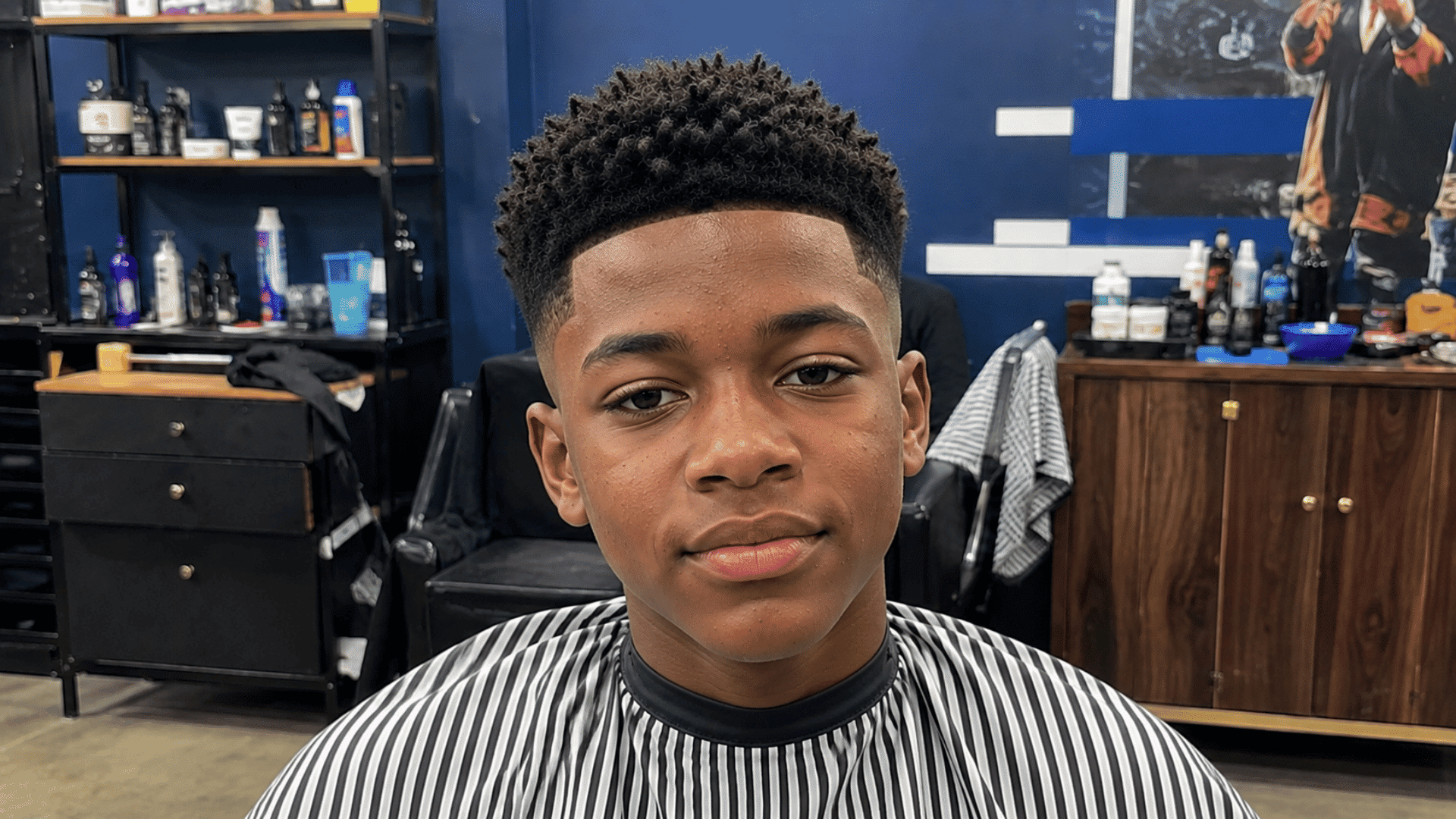 teenager sporting crew cut with mid fade haircut sitting in barber chair wearing striped cape