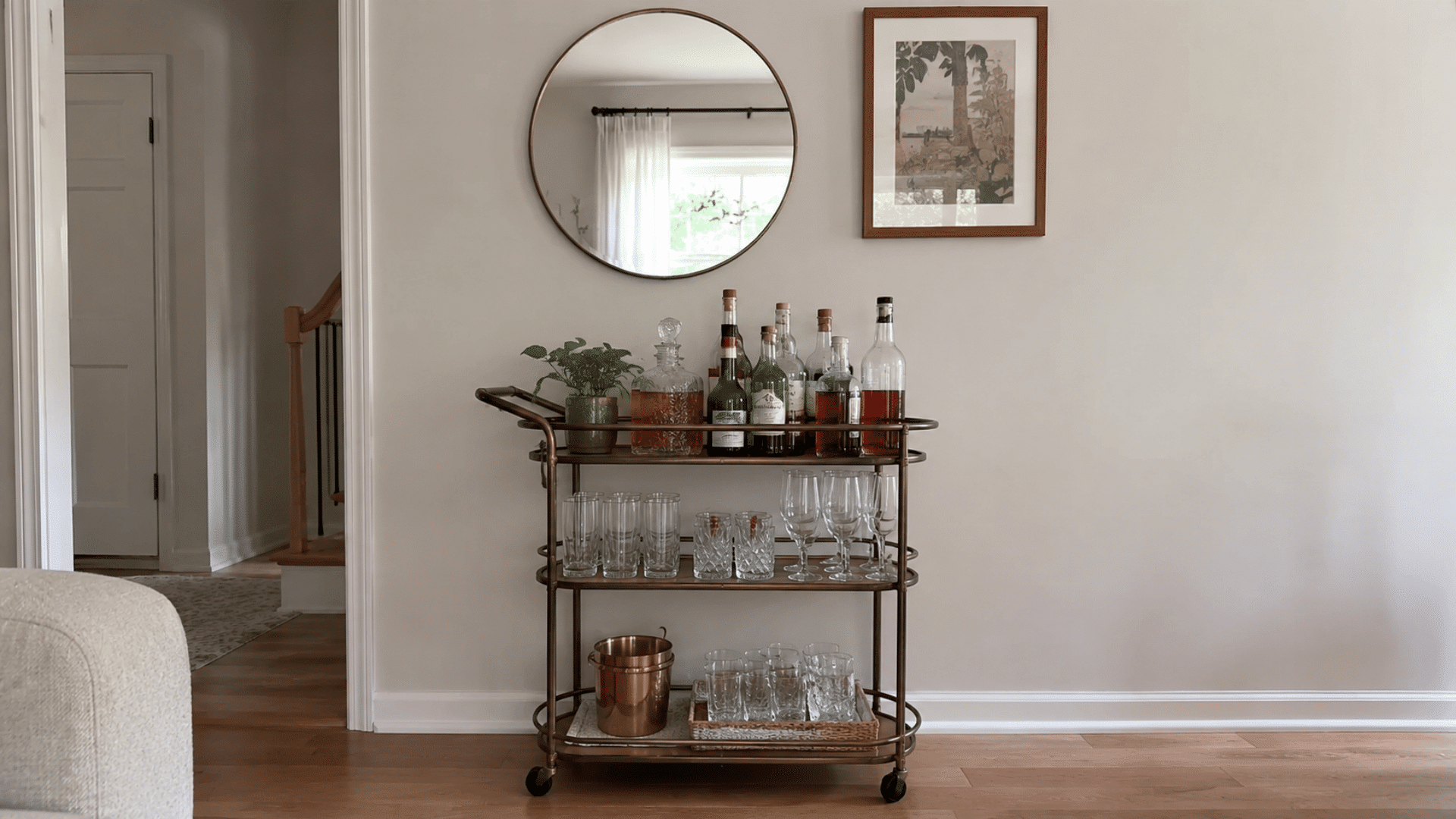 Three-tier brass bar cart with bottles, glassware, and copper ice bucket beneath a round mirror and framed print