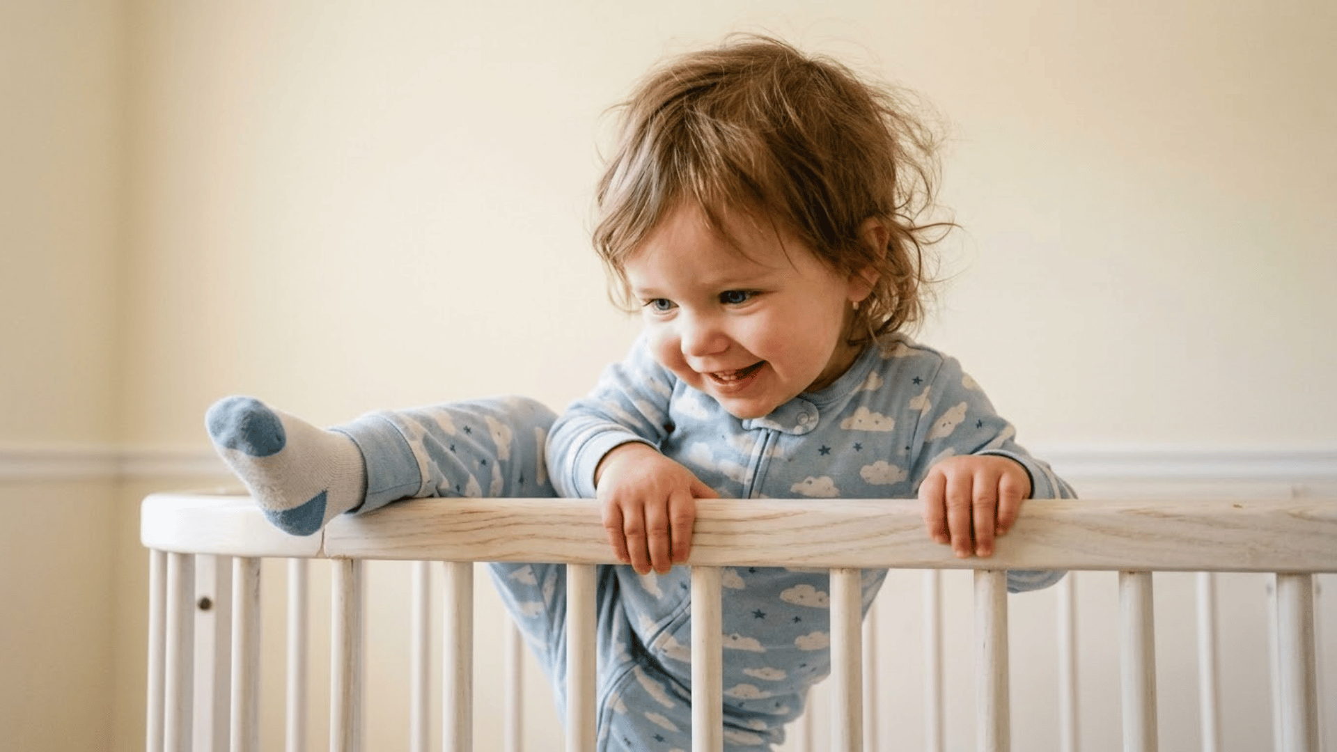 toddler in blue cloud-print pajamas smiling and climbing over the white rail of a crib, one foot swung over the side