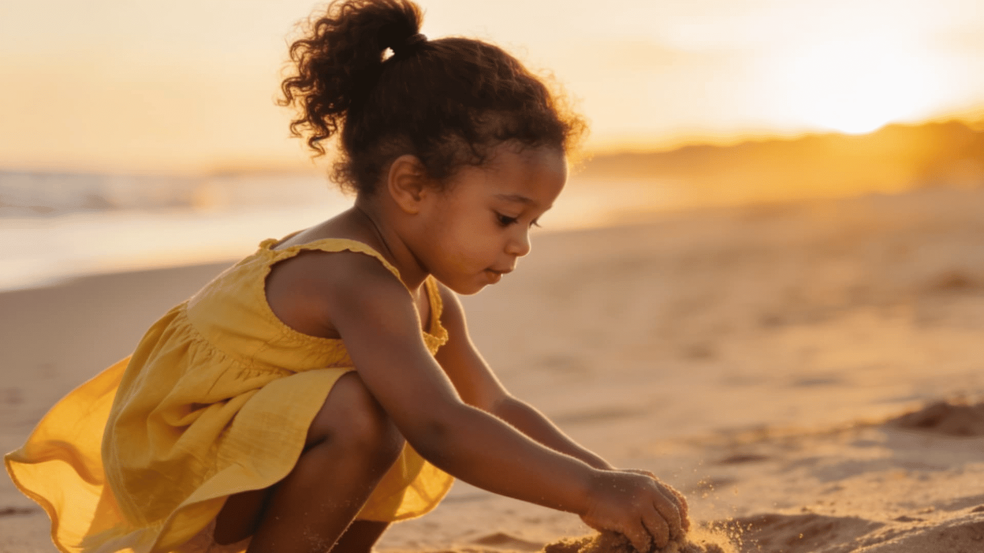 toddler girl with a high curly ponytail building a sand mound on the beach during sunset