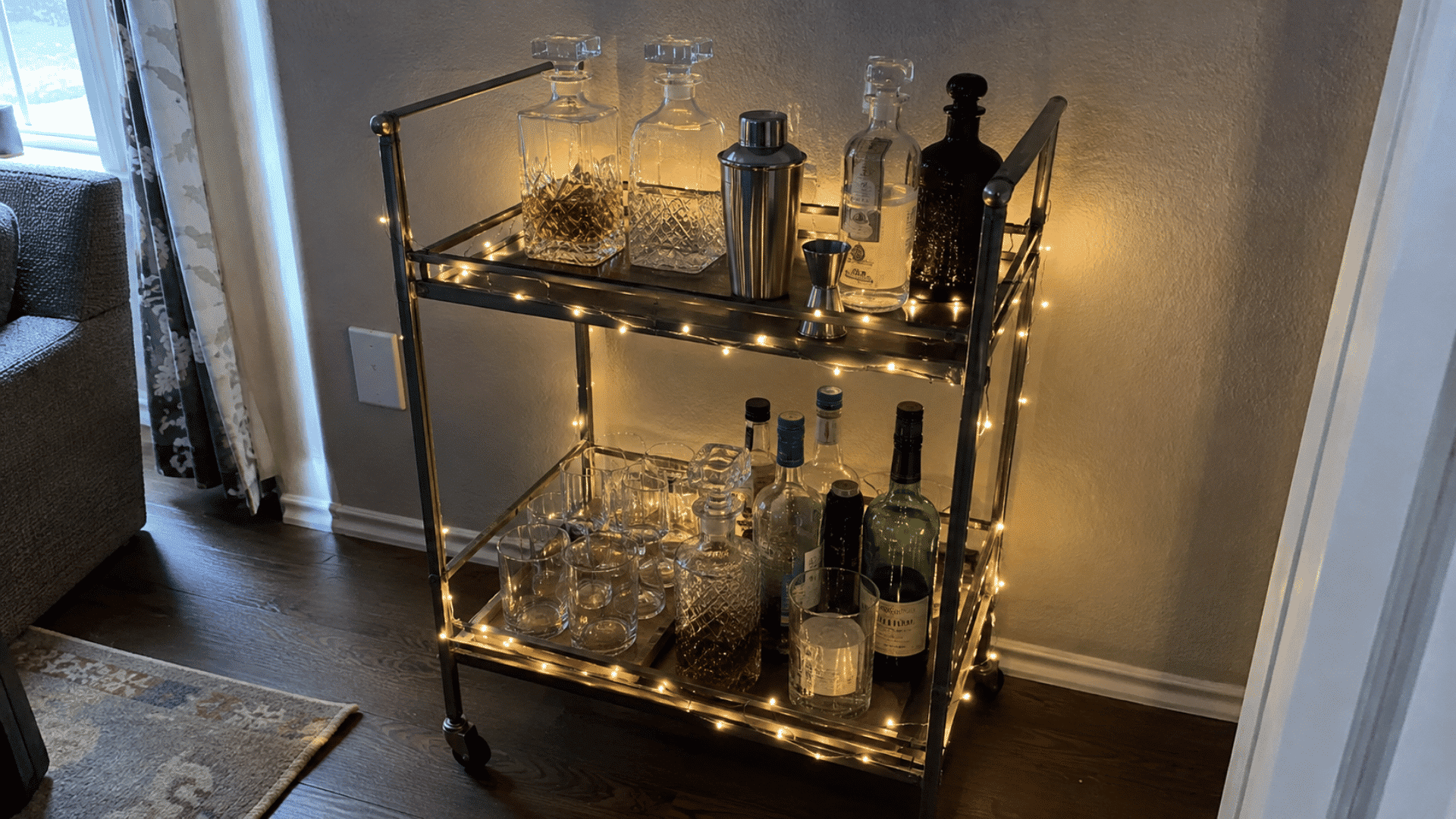 Two-tier metal bar cart wrapped in glowing string lights, holding decanters, shaker, jigger, glasses, and bottles