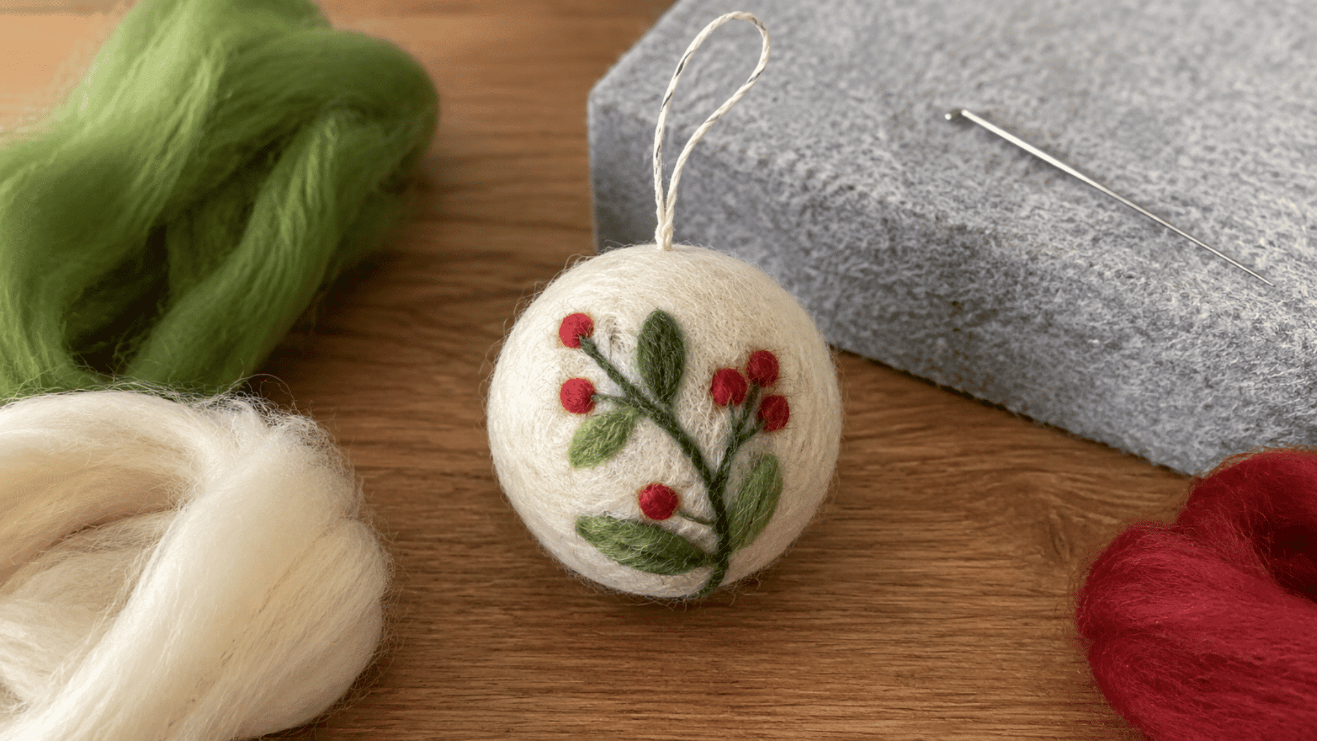White felted round ornament with holly berry design and hanging loop, surrounded by wool roving