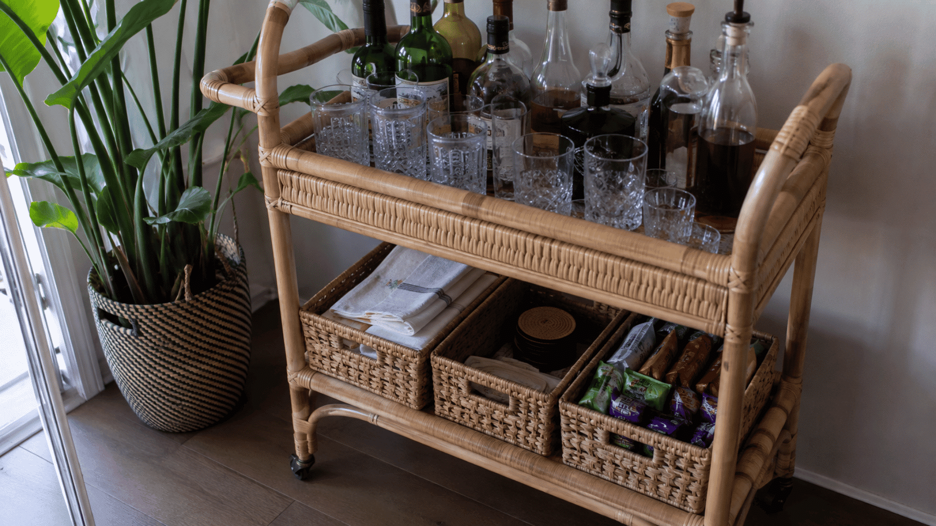 Woven rattan bar cart with wine bottles, crystal glasses, and lower baskets holding napkins, coasters, and snacks