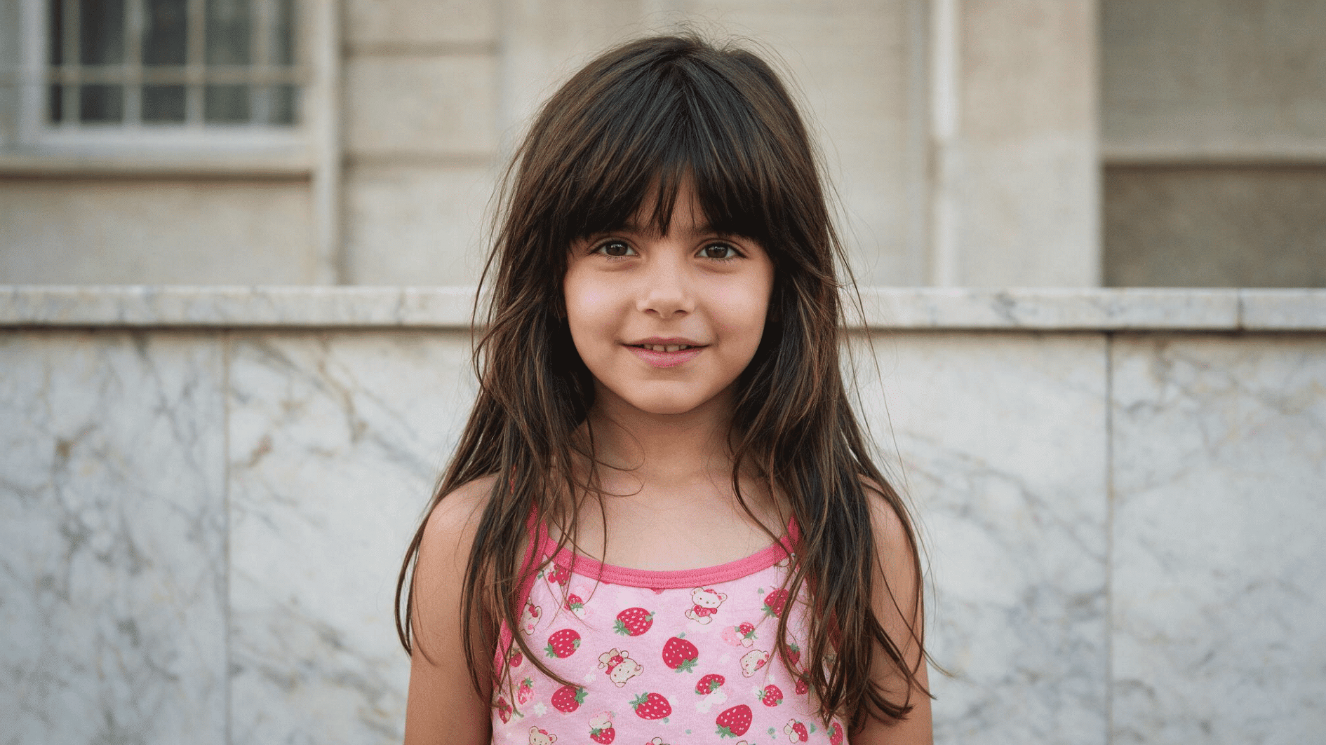 young child with long dark hair with butterfly hairstyle wearing pink top standing outdoors against a light wall background