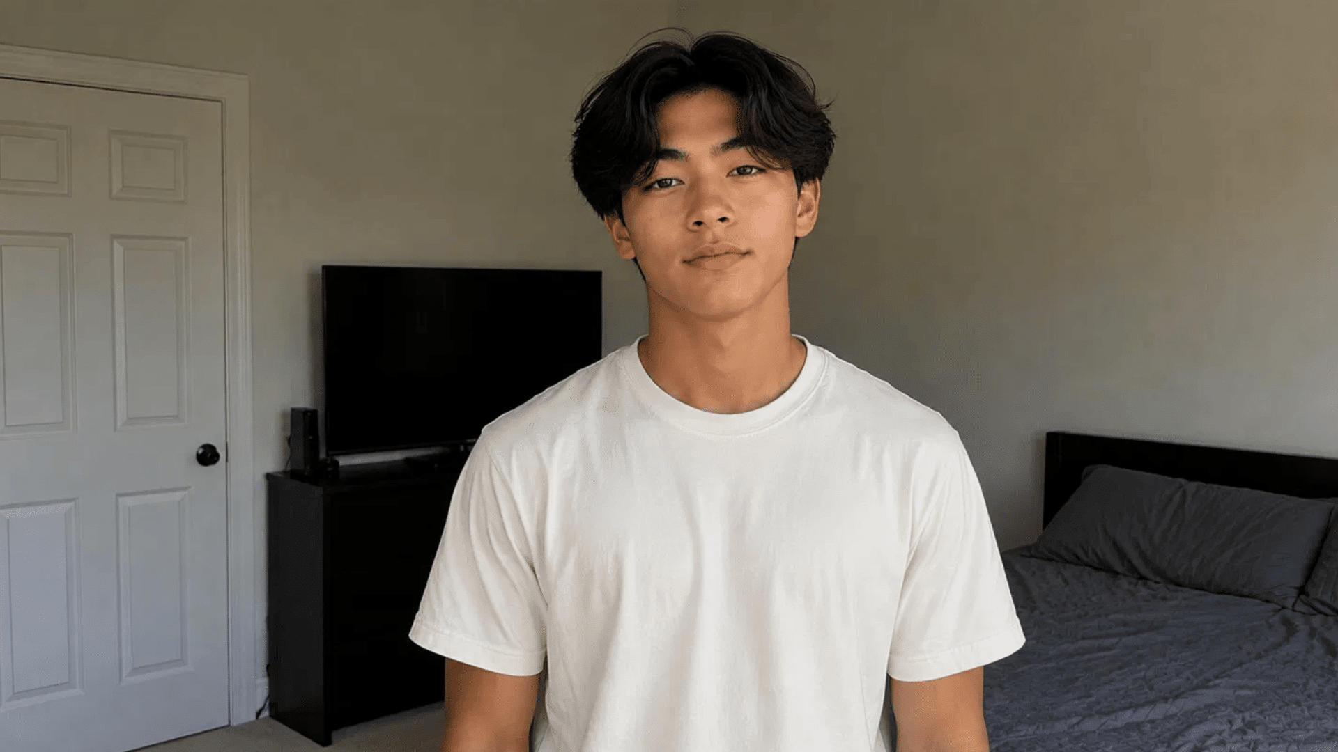 young man with a curtains hairstyle wearing a white t-shirt, standing in a bedroom with a tv and bed