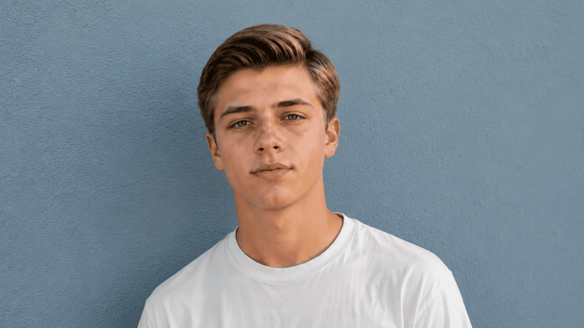 young man with a side part hairstyle wearing a white t-shirt against a dusky concrete wall
