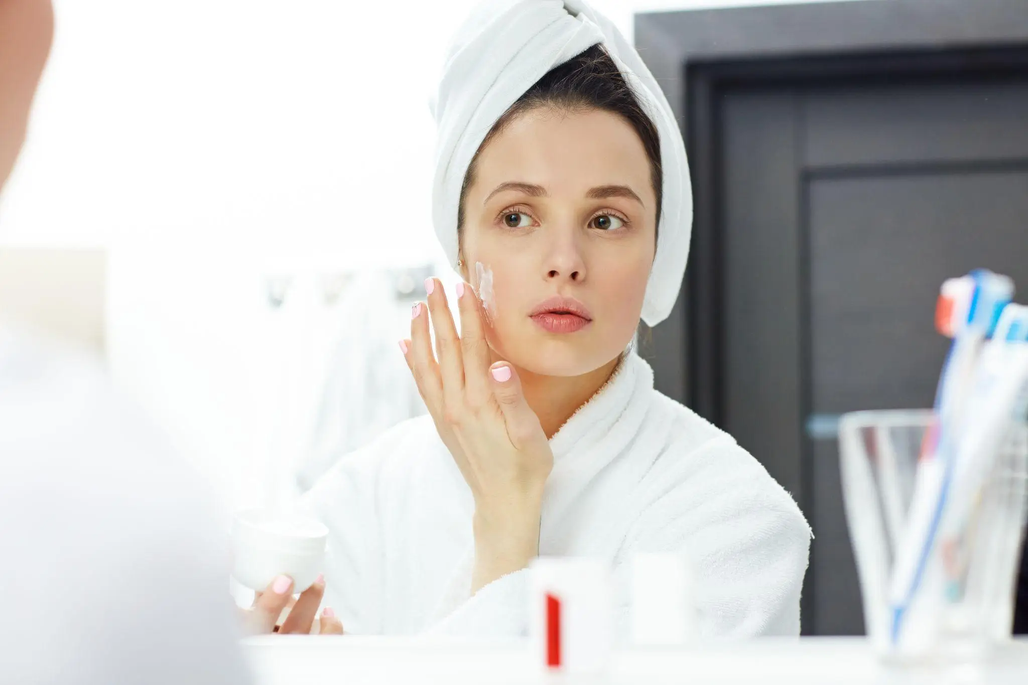 Woman applying cream to face in bright bathroom wearing white bathrobe and towel turban