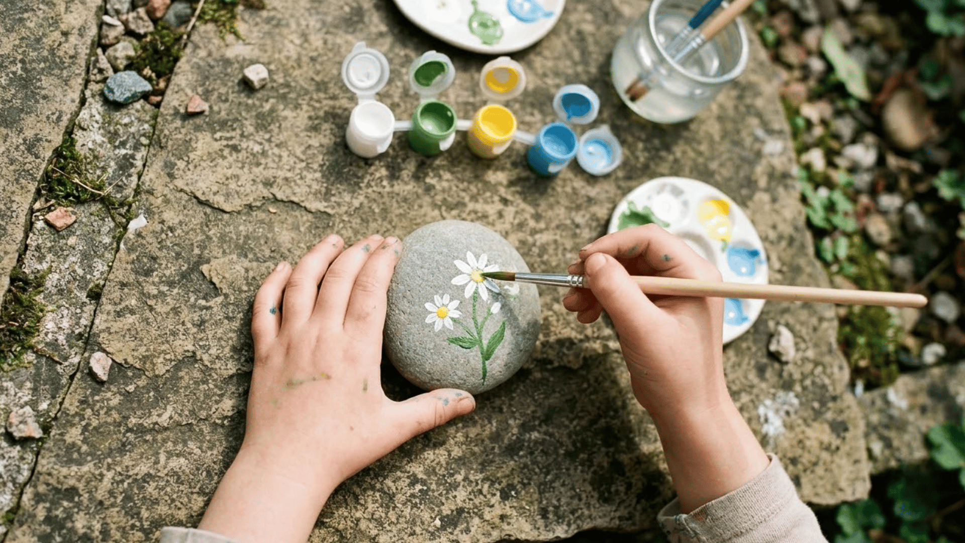 hild sitting near flower bed painting smooth rock into colorful garden plant marker