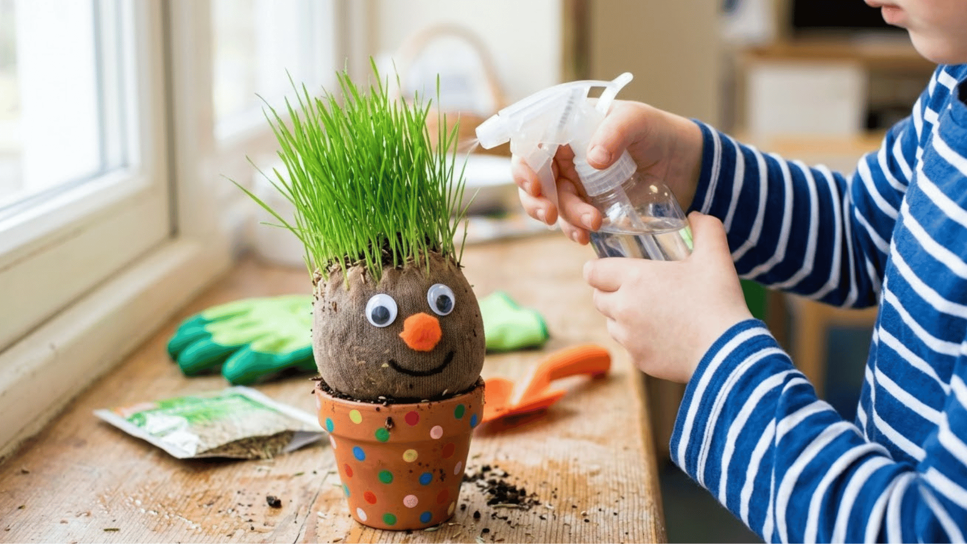 child at garden table sprinkling grass seed into decorated cup with painted face