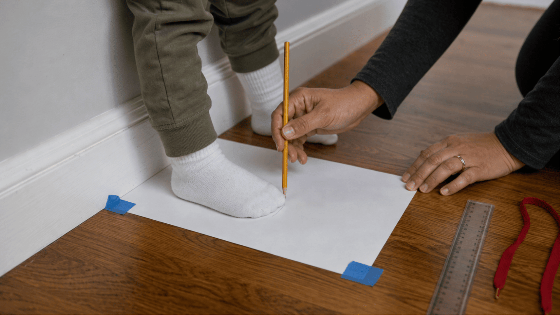 parent holding a pencil upright to mark the longest toe of a toddler standing heel against the baseboard