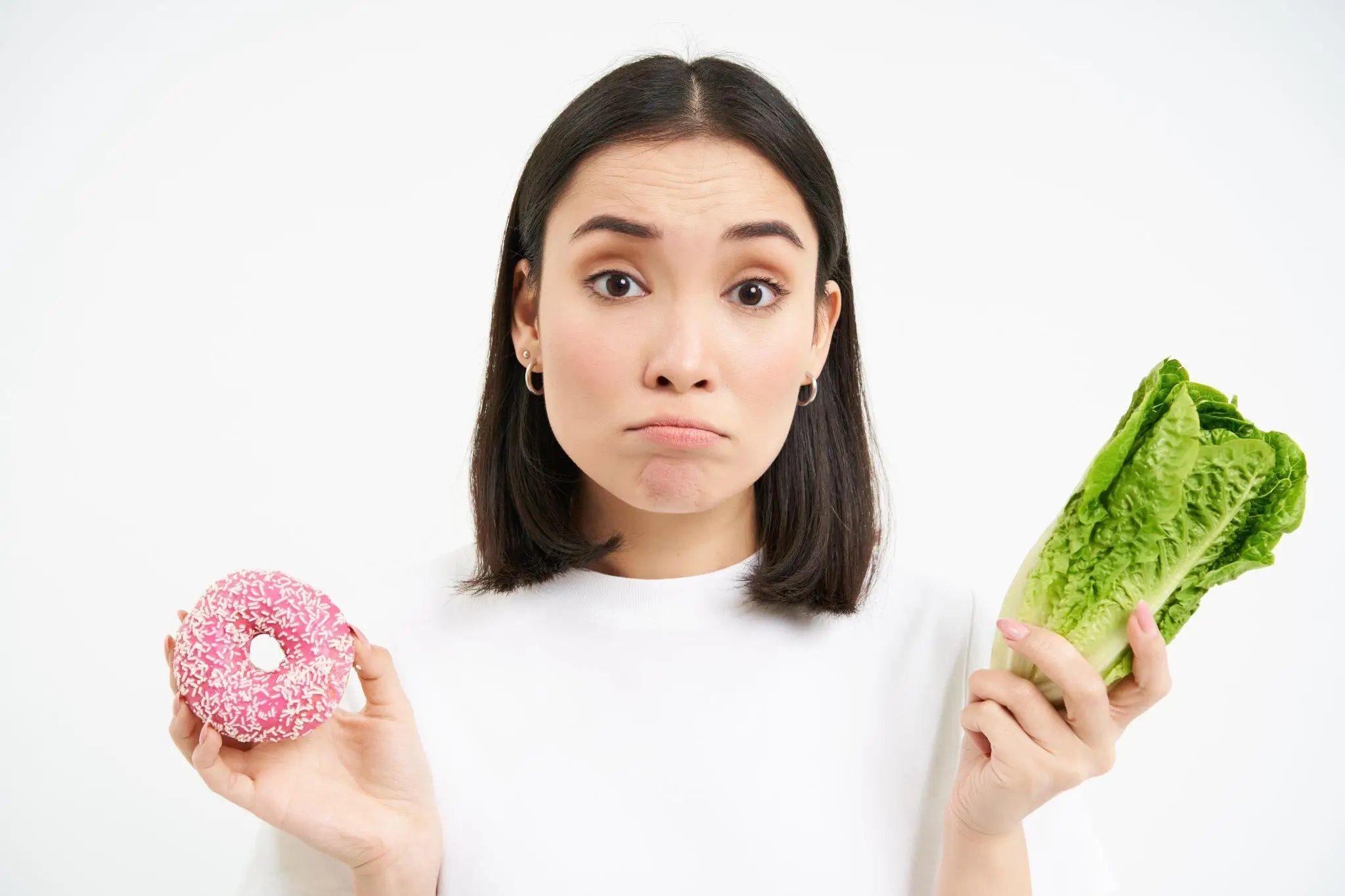Woman holding pink donut and romaine lettuce with uncertain expression against white background