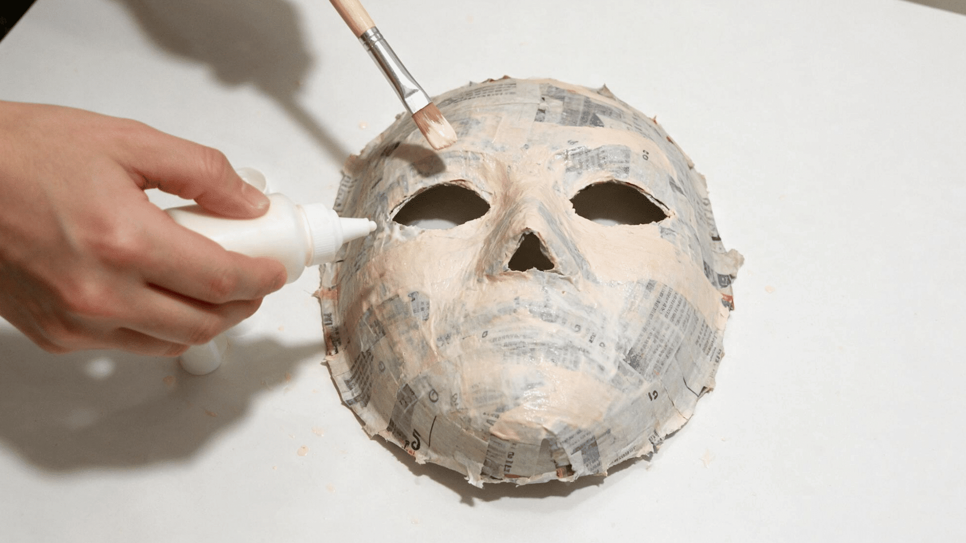 hand painting a white paper mache mask with a brush on a table