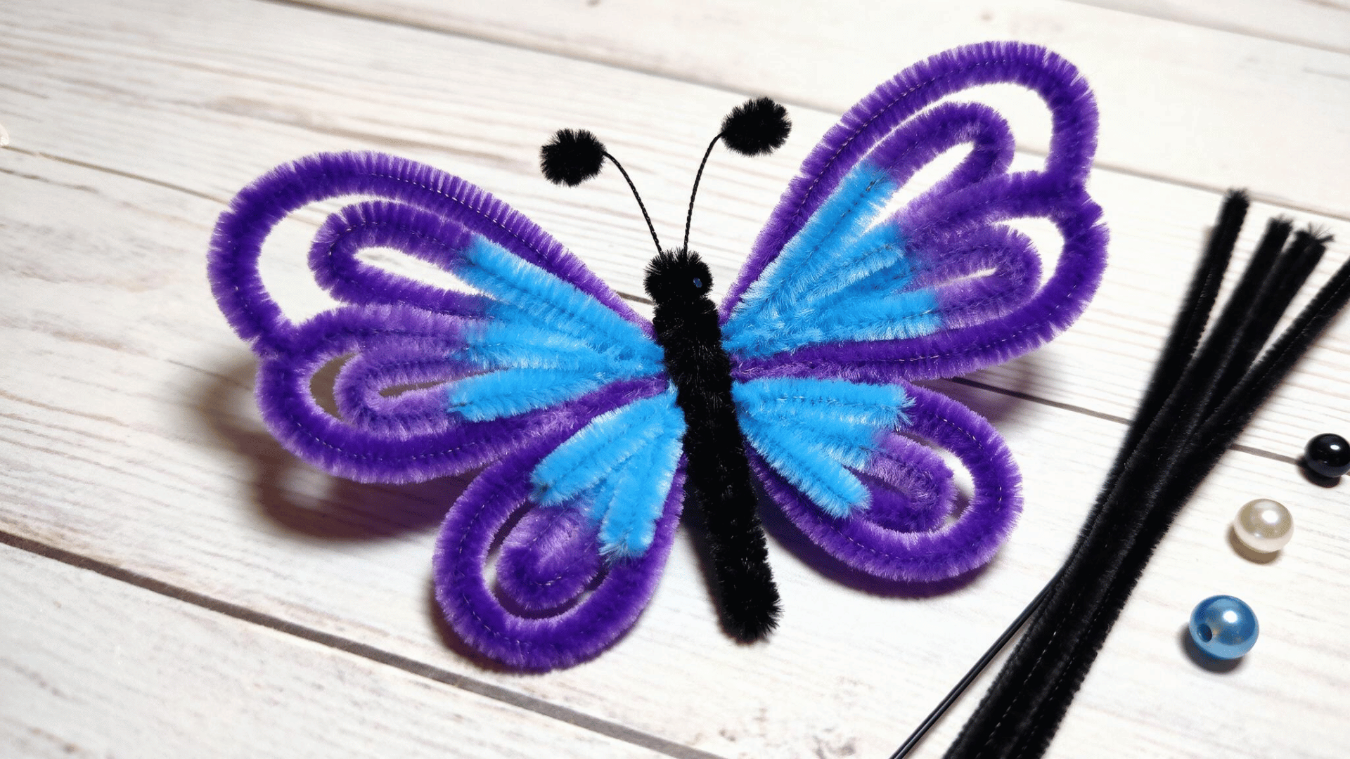 purple and blue pipe cleaner butterfly with looped wings black body and antennae resting on a white wood table