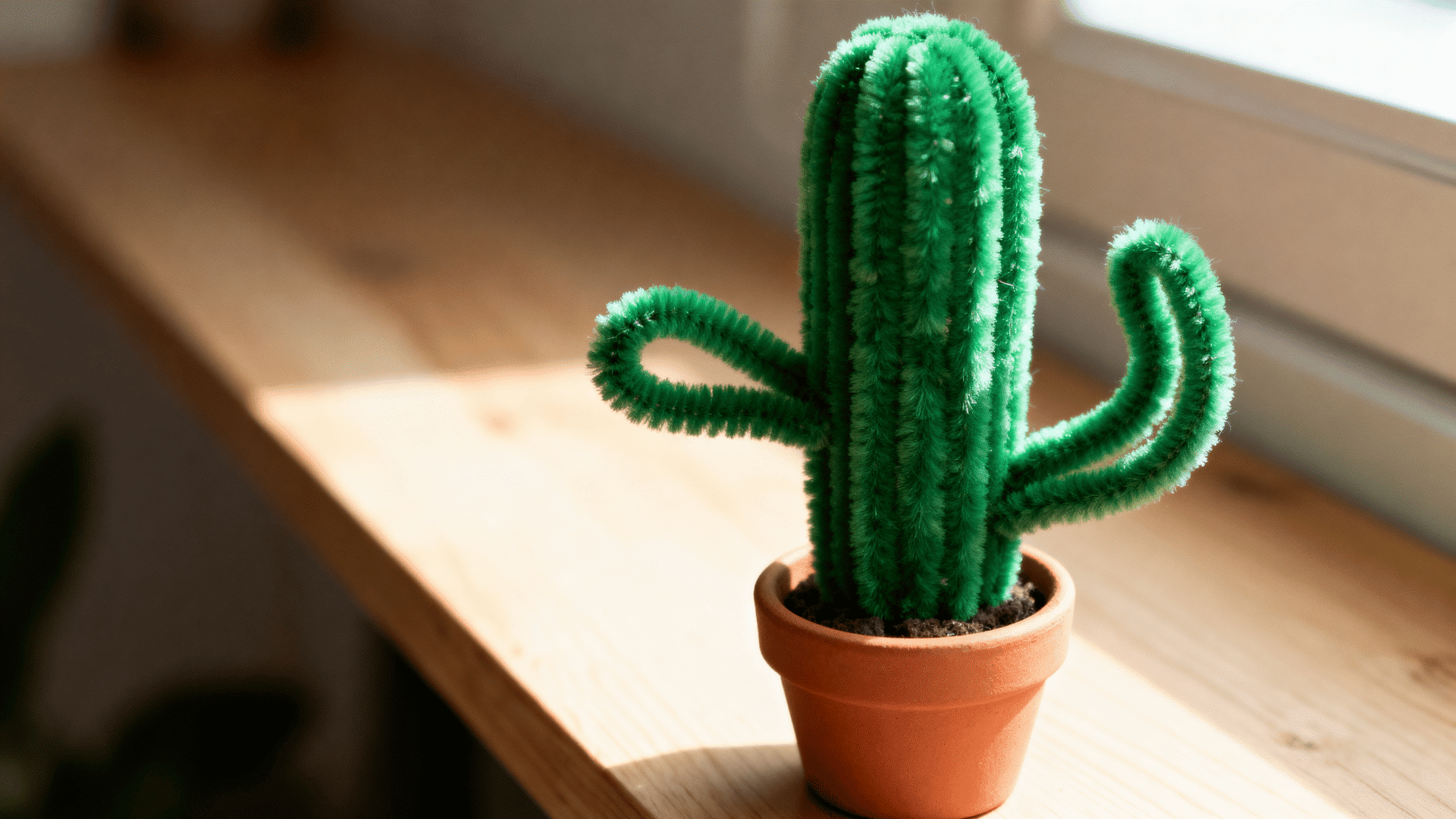 green pipe cleaner cactus with two curved arms standing in a small terracotta pot on a sunny wooden windowsill