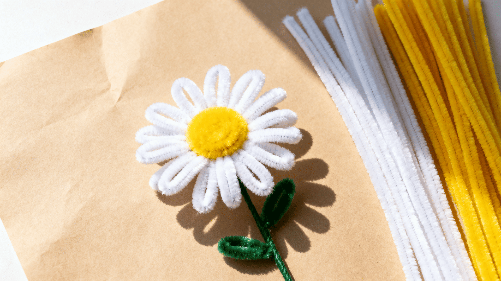 white pipe cleaner daisy with yellow center and green stem resting on kraft paper beside loose chenille stems nearby