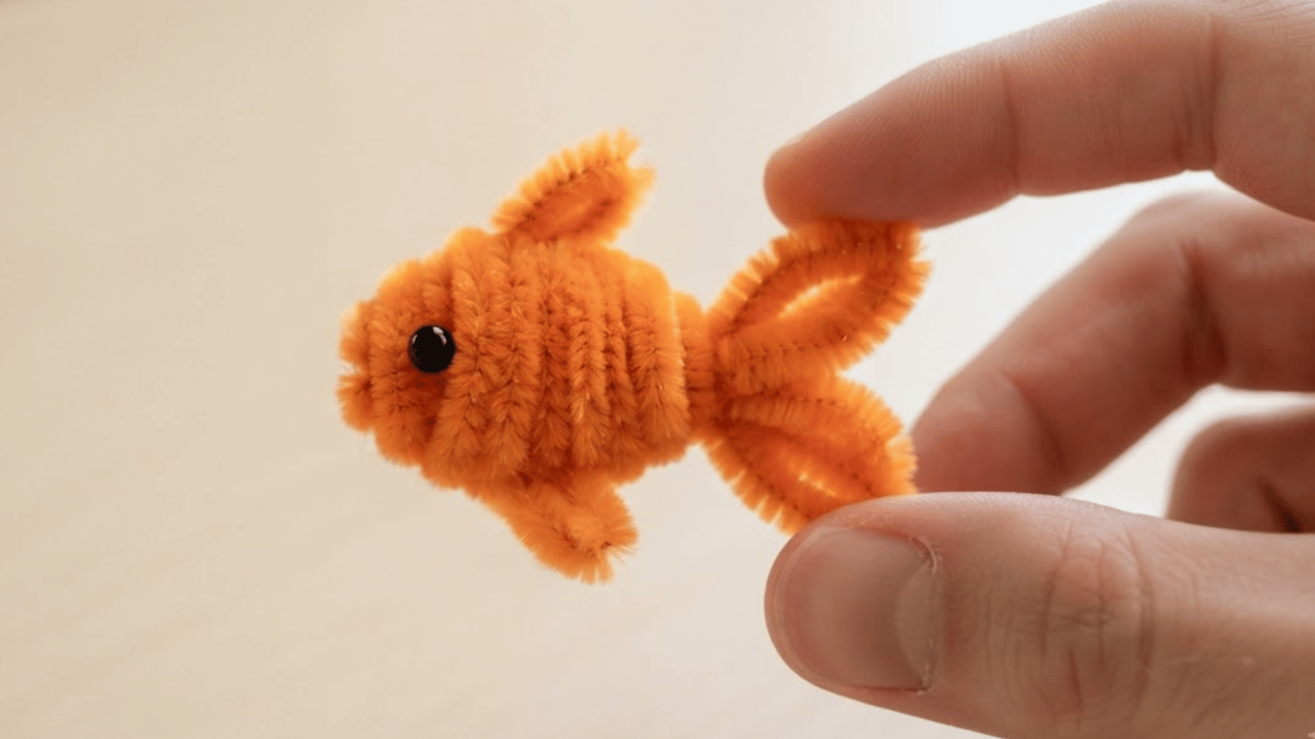 orange pipe cleaner fish with rounded body small fins and looped tail held gently between two fingertips in daylight