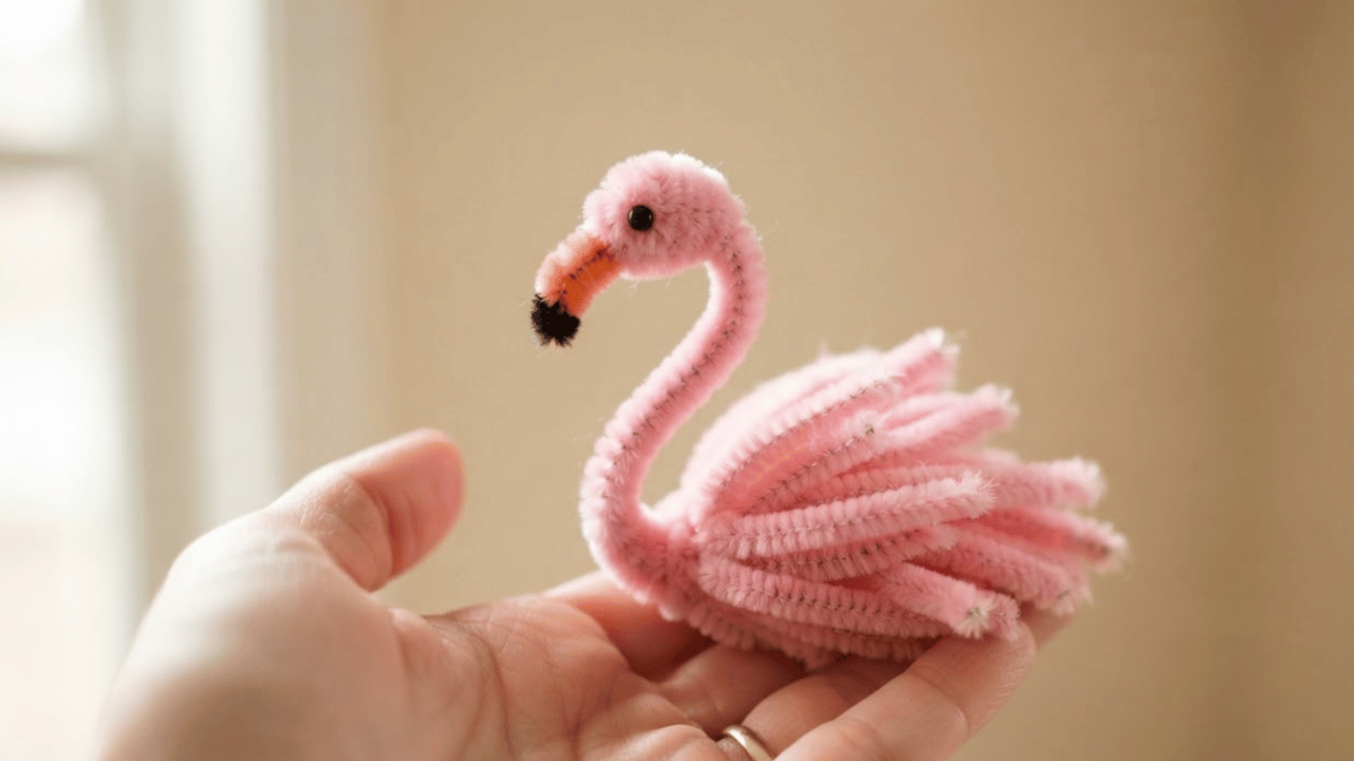 pink pipe cleaner flamingo with curved neck layered feather-like body and black-tipped beak resting on a hand indoors