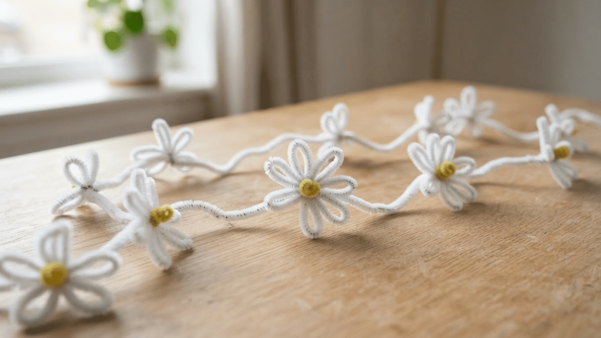 white pipe cleaner daisy garland with yellow centers arranged loosely across a wooden table near a bright window