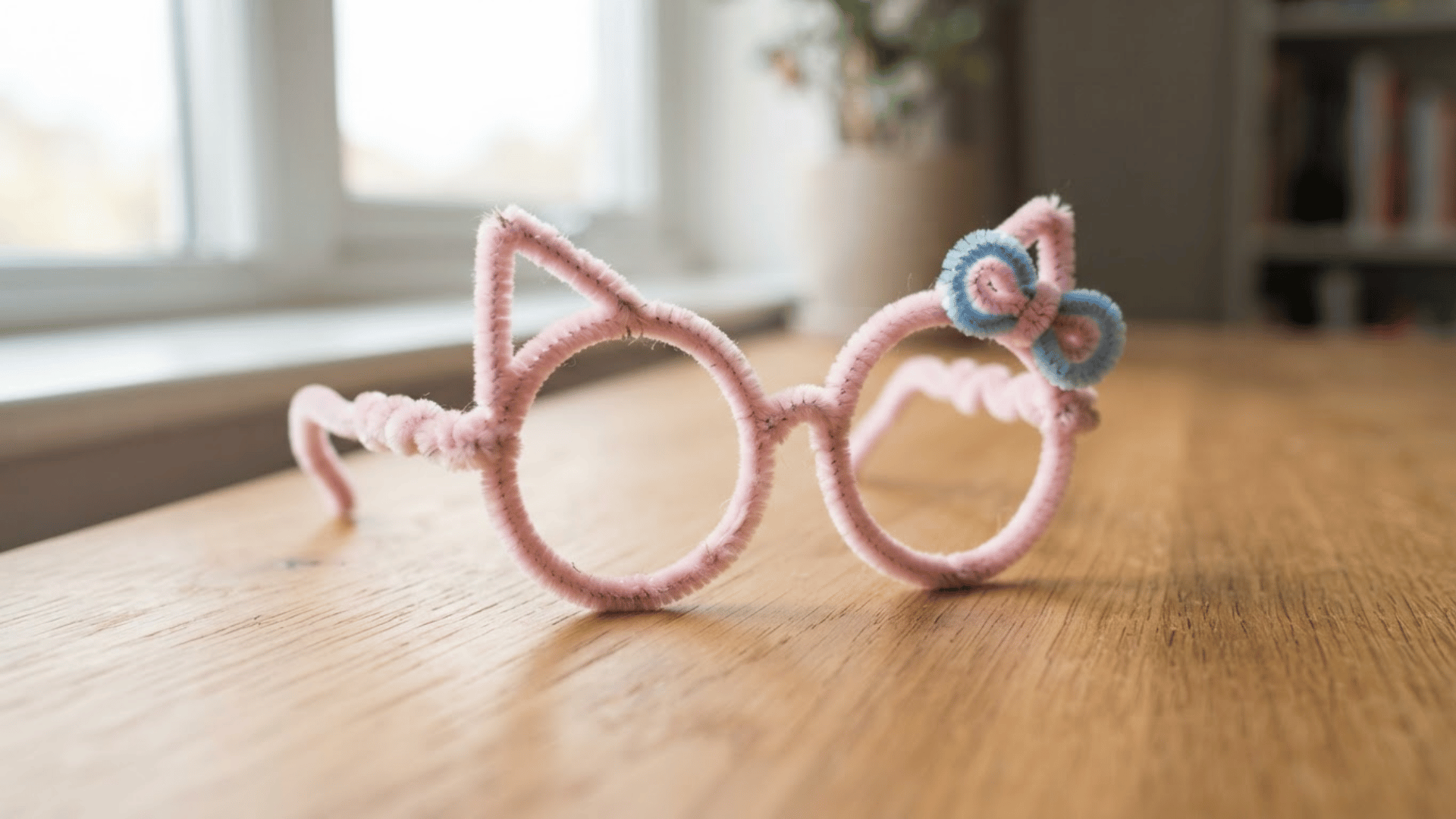 pink cat shaped pipe cleaner glasses with round frames pointed ears and a blue bow resting on a wooden table indoors