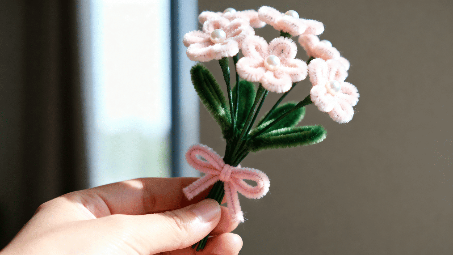 pink pipe cleaner flower bouquet with pearl centers green stems leaves and a matching bow held gently near a window