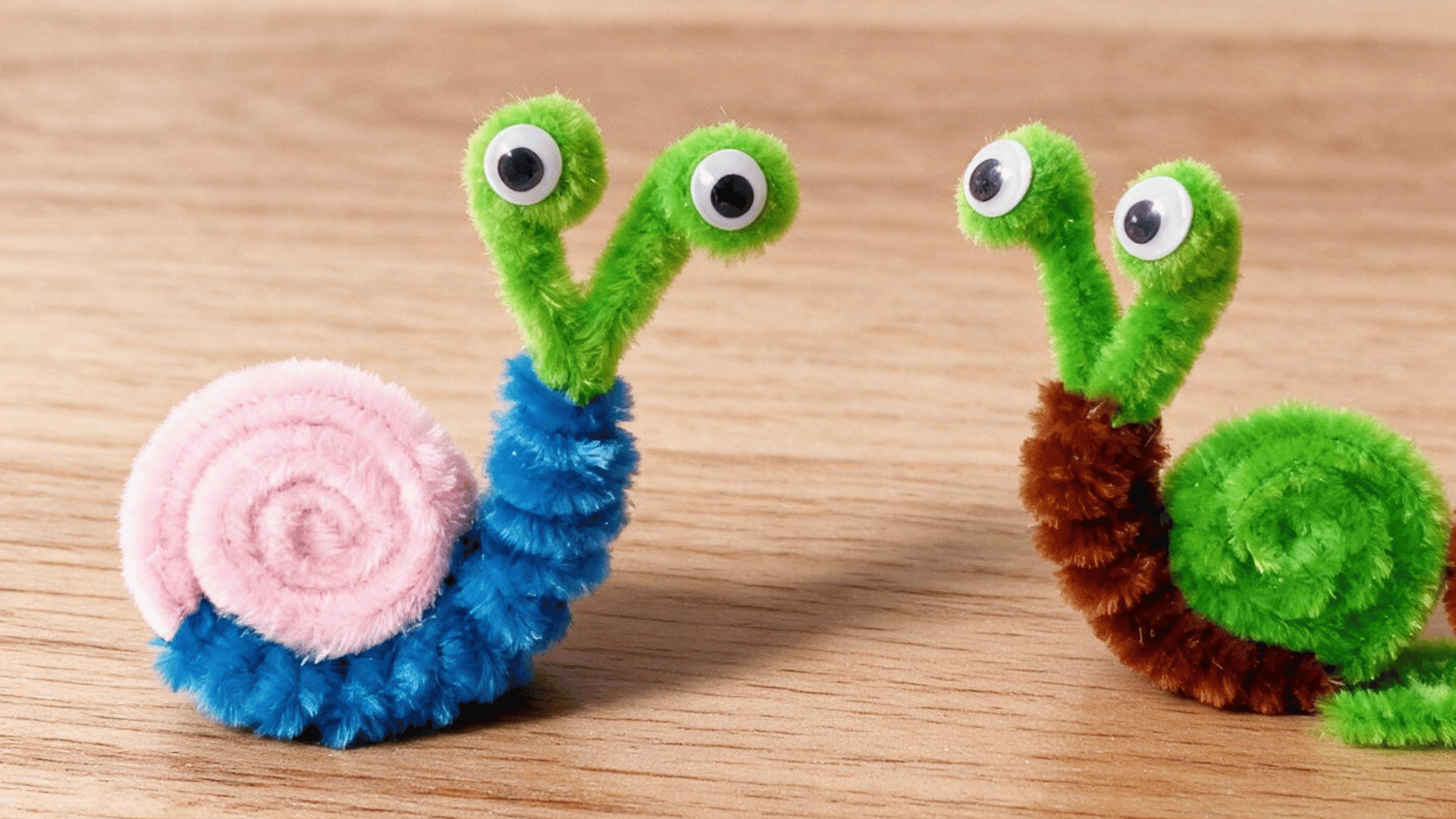 two colorful pipe cleaner snails with spiral shells and googly eyes standing on a light wooden tabletop surface