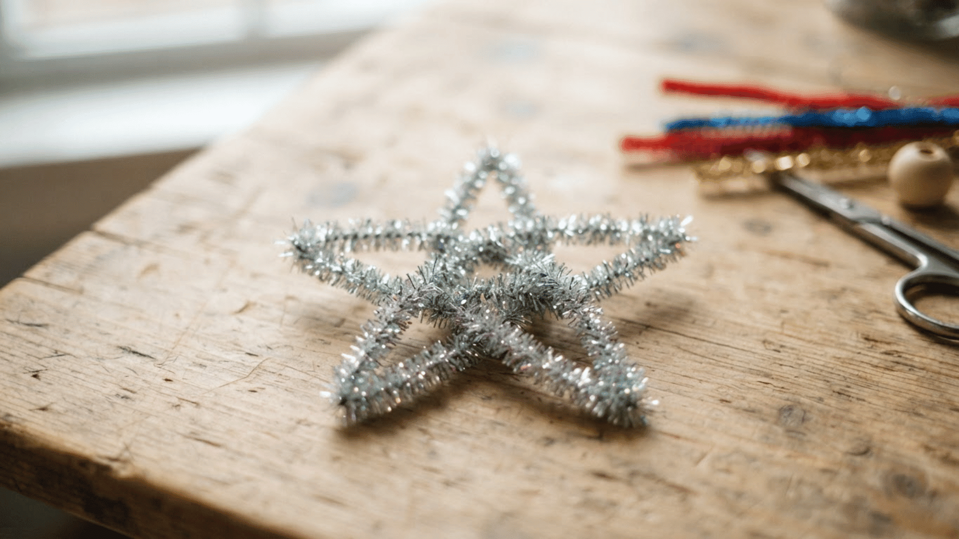silver metallic pipe cleaner star with five points resting on a rustic wooden table beside a few craft supplies