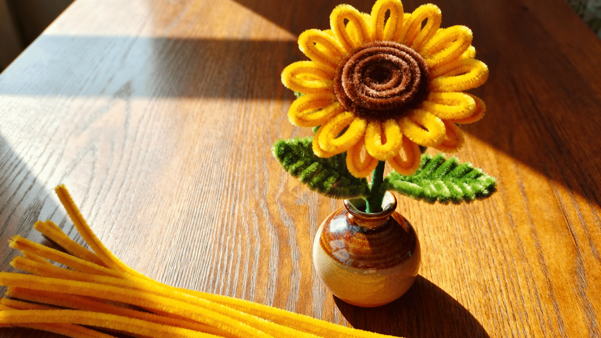 yellow pipe cleaner sunflower with brown spiral center and green leaves arranged in a small vase on a wood table