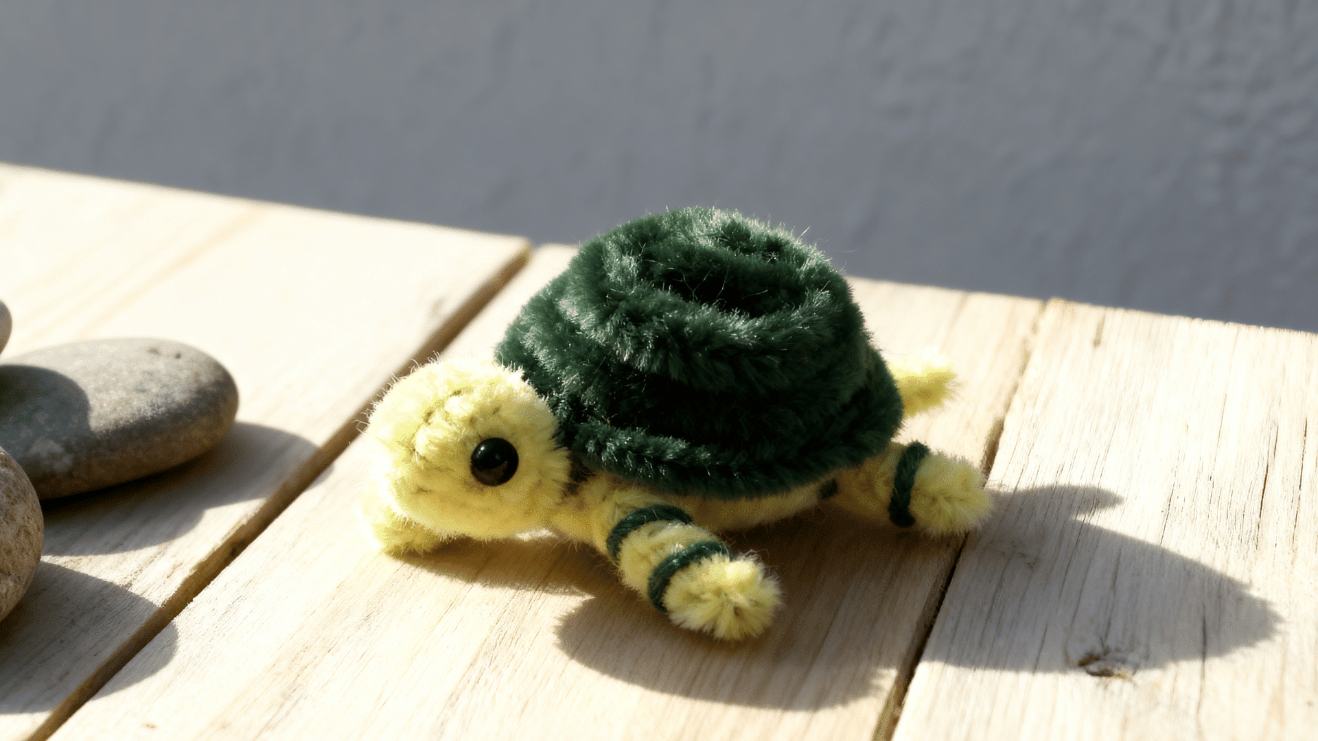green pipe cleaner turtle with dark spiral shell short legs and black eyes resting on a pale wooden tabletop indoors