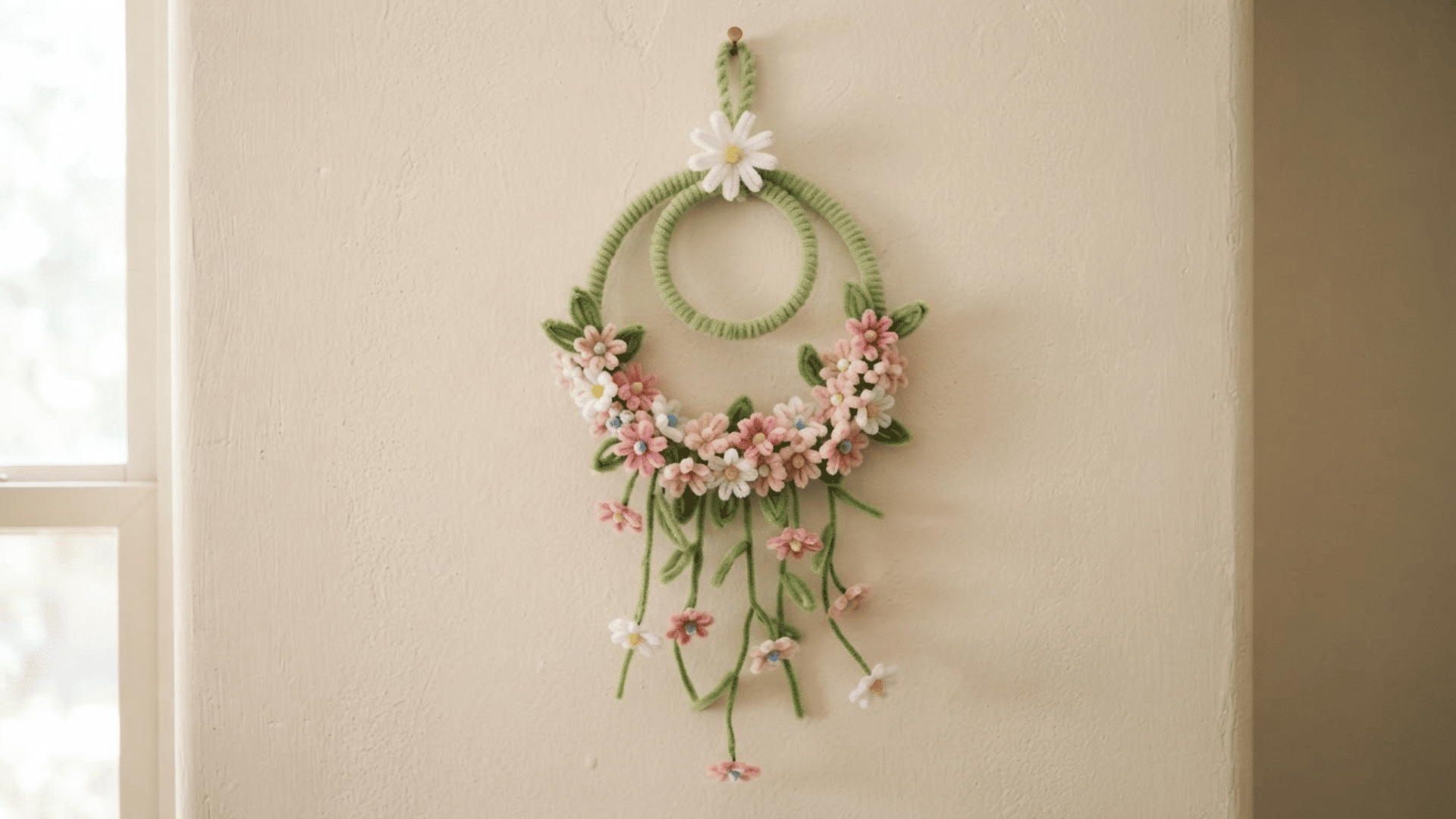 green pipe cleaner floral wall hanging with pink and white flowers and dangling stems displayed on a cream wall indoors