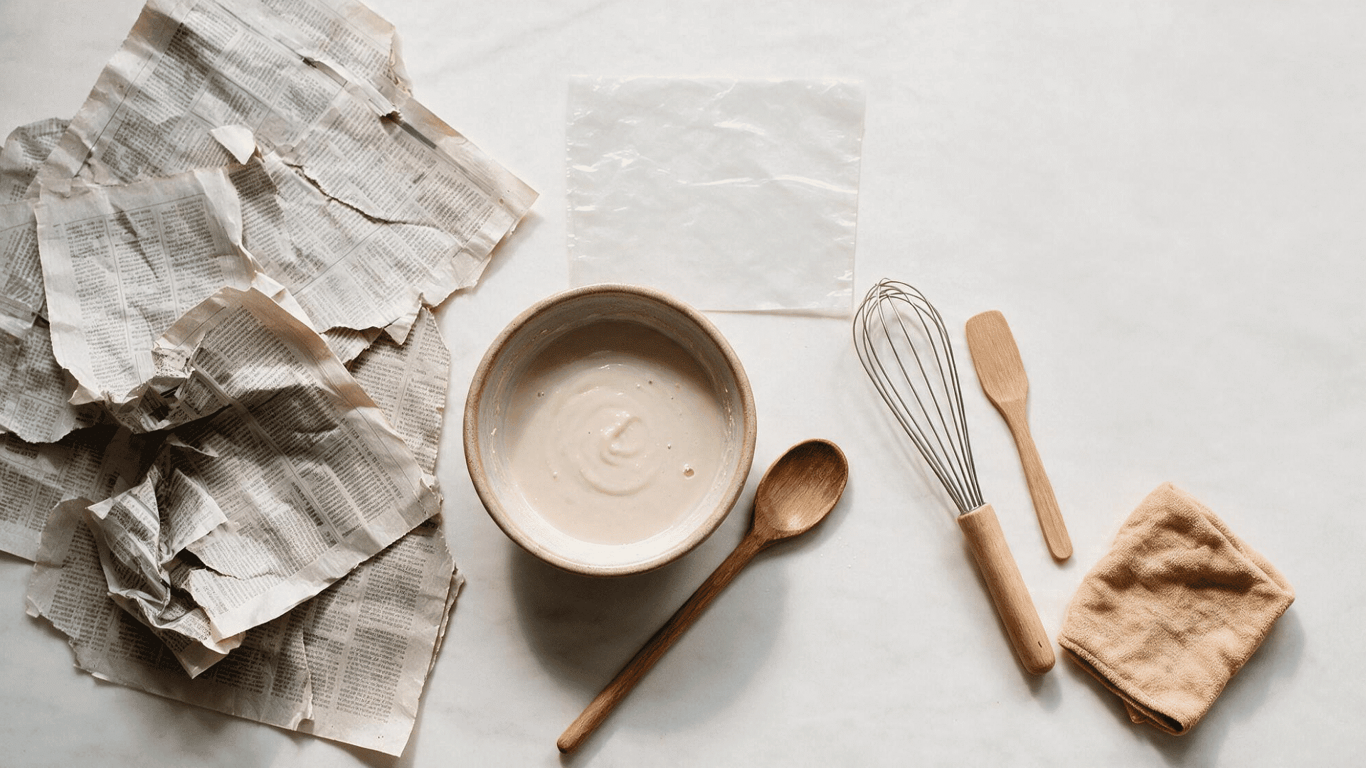 flat lay of paper mache supplies including newspaper, flour, water, whisk, and wooden spoon prepared before starting